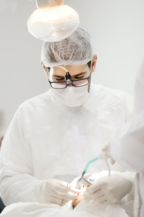 A dentist wearing a white coat, hair net, mask, gloves, and magnifying glasses works on a patient's teeth using dental instruments, illuminated by a dental overhead light.