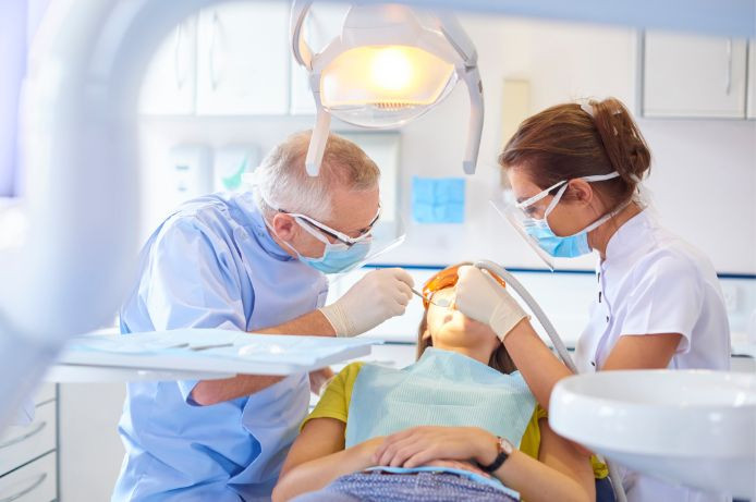 A dentist and dental assistant, both wearing masks and protective eyewear, perform a dental procedure on a patient lying in a dental chair.