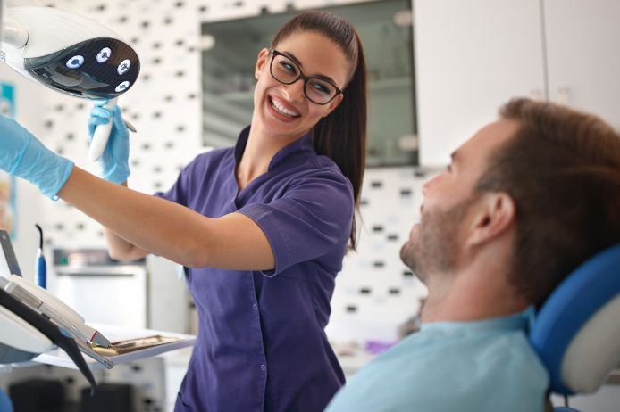 A female dentist wearing glasses and a purple uniform smiles while assisting a male patient who is seated and smiling in a dental chair.