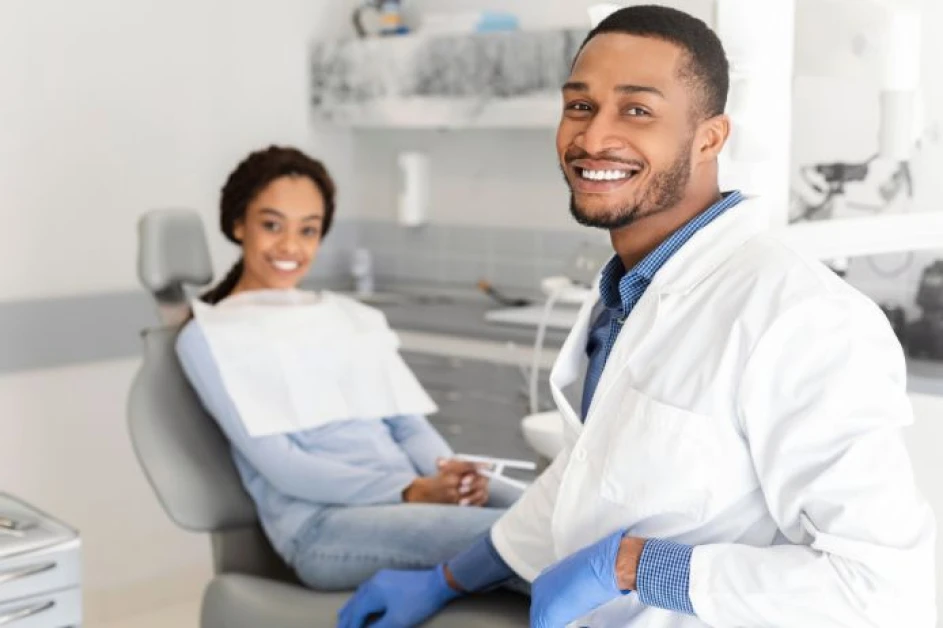  A smiling NHS dentist in a white coat and blue gloves sits on a stool in a well-equipped dental practice